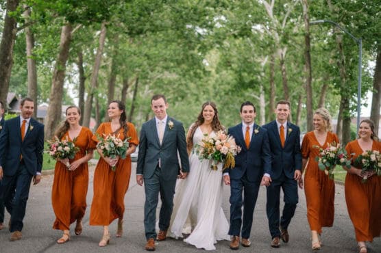 A bride and groom walk hand in hand down a tree-lined street, smiling and surrounded by three bridesmaids in rust-orange dresses and three groomsmen in navy suits with orange ties, all holding bouquets. by Justin Salem Meyer