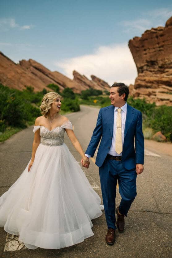 A bride in a white gown and a groom in a blue suit walk hand in hand down a scenic road, smiling at each other, with rocky red formations and greenery in the background. by Justin Salem Meyer