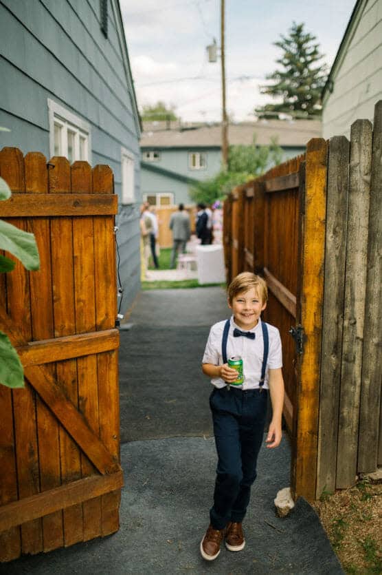 A smiling young boy in suspenders, a bow tie, and dress shoes stands by an open wooden gate, holding a can of soda, with a gathering of people visible in the yard behind him. by Justin Salem Meyer