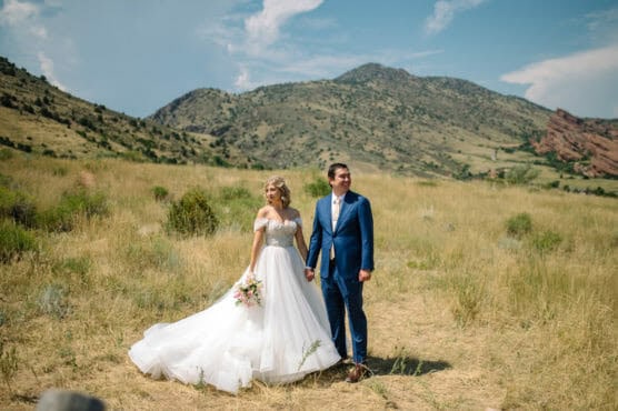 A bride in a white gown and a groom in a blue suit stand holding hands in a grassy field with mountains in the background under a blue sky. by Justin Salem Meyer