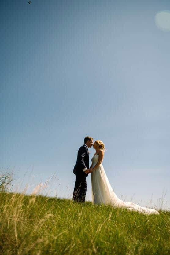 A bride and groom stand on a grassy hill, holding hands and sharing a kiss under a clear blue sky, with sunlight highlighting the bride’s white dress. by Justin Salem Meyer