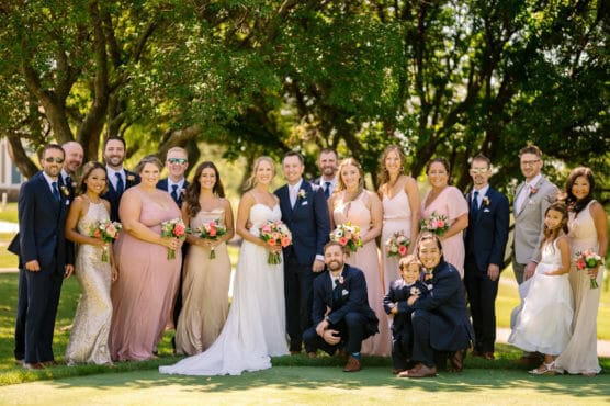 A wedding party poses outdoors on a sunny day. The bride, groom, bridesmaids in pink dresses, and groomsmen in suits stand together, all smiling, holding bouquets, and surrounded by green trees. by Justin Salem Meyer