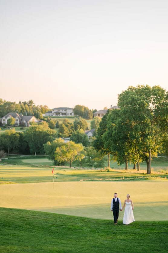 A bride and groom walk hand in hand across a sunlit golf course, surrounded by green grass, large trees, and distant houses under a clear sky. by Justin Salem Meyer