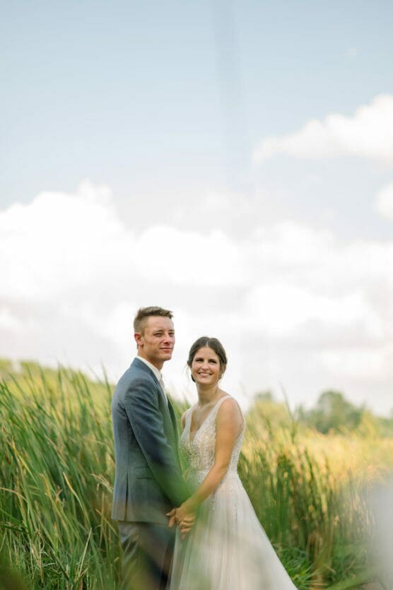 A bride and groom stand holding hands in a field of tall grass, smiling at the camera under a bright, partly cloudy sky. by Justin Salem Meyer