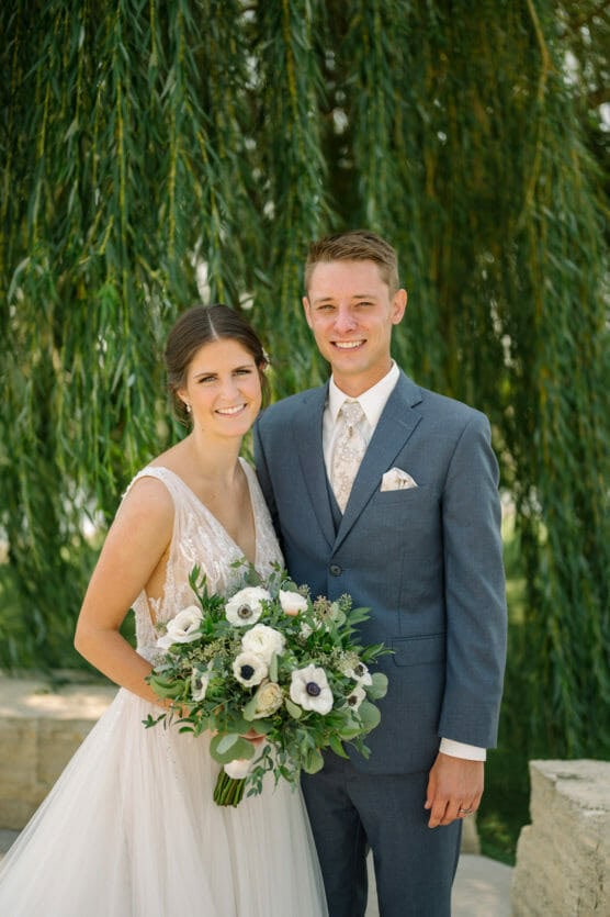 A bride in a white dress holding a bouquet of white flowers stands next to a groom in a blue suit and tie. They are smiling outdoors in front of lush, green hanging willow branches. by Justin Salem Meyer