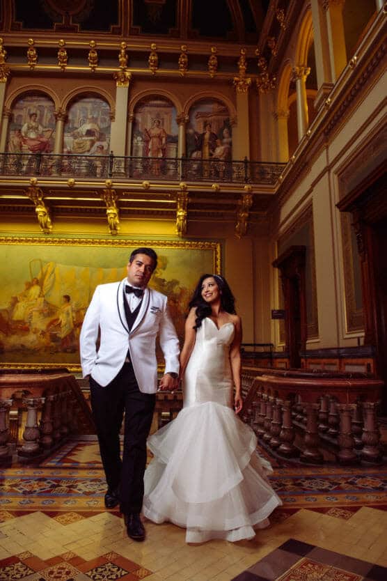 A bride and groom walk hand in hand inside an ornate hall with classical paintings, high ceilings, and decorative columns. The groom wears a white tuxedo and the bride wears a strapless white gown with a flared skirt. by Justin Salem Meyer