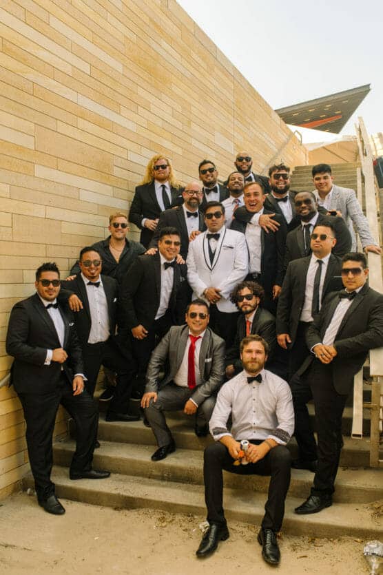 A group of men dressed in suits and tuxedos pose together on outdoor stairs against a beige brick wall, most wearing sunglasses and smiling for the camera. by Justin Salem Meyer