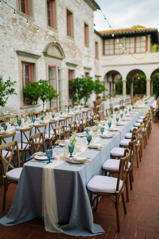 Elegant outdoor dining setup in a courtyard with long tables, white and blue tablecloths, blue glassware, and wooden chairs. String lights are hung above, and the setting is beside a white brick building with arched windows. by Justin Salem Meyer