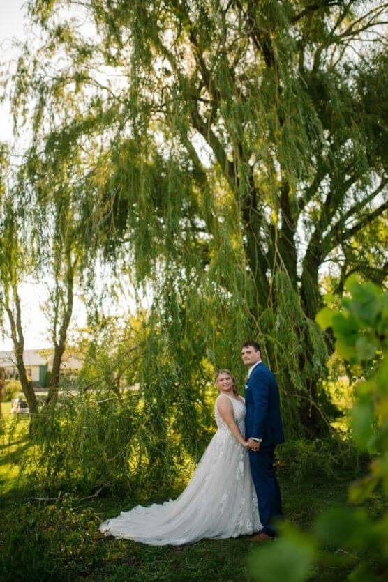 A bride and groom stand hand in hand outdoors under a large, leafy tree. The bride wears a white gown with a long train, and the groom wears a blue suit. Sunlight filters through the branches, creating a serene, romantic scene. by Justin Salem Meyer