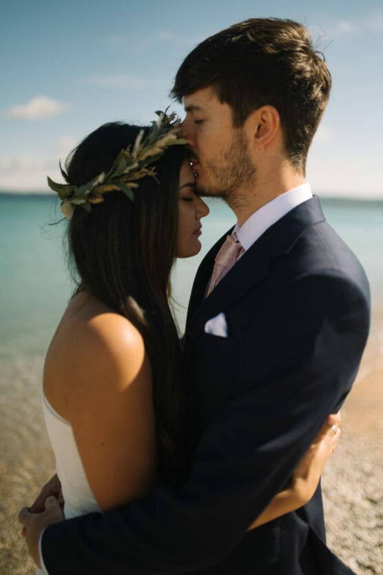 A couple embraces on a beach; the man in a suit kisses the woman’s forehead. The woman, wearing a white dress and a leafy crown, closes her eyes. The ocean and sky are blurred in the background. by Justin Salem Meyer