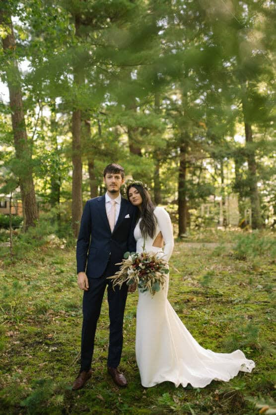 A bride in a long white dress holding a bouquet stands closely with a groom in a dark blue suit and tie in a forest with sunlight filtering through green trees. by Justin Salem Meyer