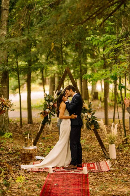 A bride and groom kiss during an outdoor wedding ceremony in a forest, standing on a red rug in front of a triangular wooden arch decorated with flowers and pampas grass. Trees and greenery surround them. by Justin Salem Meyer