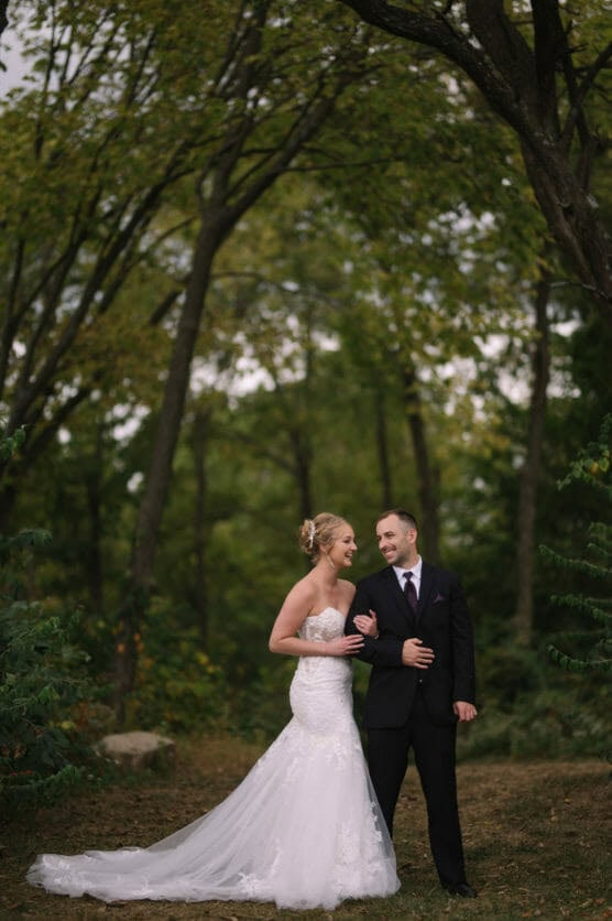 A bride in a strapless white gown and a groom in a black suit stand together outdoors, smiling at each other under tall, leafy trees in a forest setting. by Justin Salem Meyer