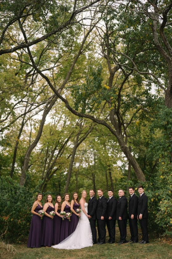 A wedding party poses outdoors under tall, leafy trees. The bride stands in the center with bridesmaids in purple dresses on her left and groomsmen in black suits on her right, all smiling for the photo. by Justin Salem Meyer
