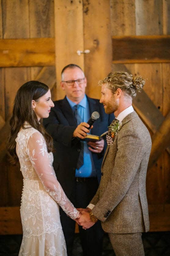 A bride and groom hold hands and face each other during their wedding ceremony. An officiant stands behind them, holding a microphone and a notebook, with rustic wooden doors in the background. by Justin Salem Meyer