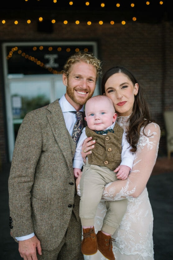 A smiling couple in formal attire pose together, holding a baby dressed in a vest and bow tie. The woman wears a white lace dress, and the man wears a brown suit. String lights and a brick wall are in the background. by Justin Salem Meyer