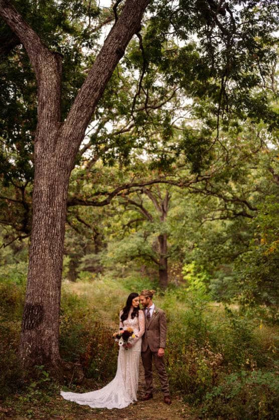 A bride in a white gown holding a bouquet stands beside a groom in a tan suit under a large tree in a lush, green forest. The couple appears peaceful and close, surrounded by natural scenery. by Justin Salem Meyer