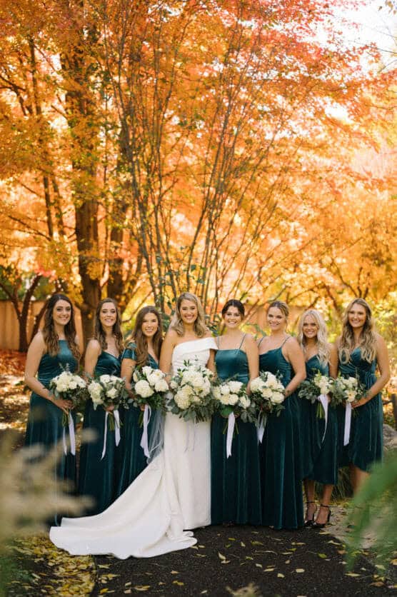 A bride in a white gown stands outdoors with six bridesmaids in teal dresses, all holding white flower bouquets. Vibrant autumn trees with orange and yellow leaves fill the background. by Justin Salem Meyer