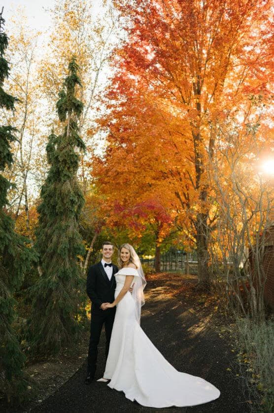 A bride in a white gown and a groom in a black suit stand holding hands on a path, surrounded by autumn trees with vibrant orange and yellow leaves, sunlight streaming through the branches. by Justin Salem Meyer