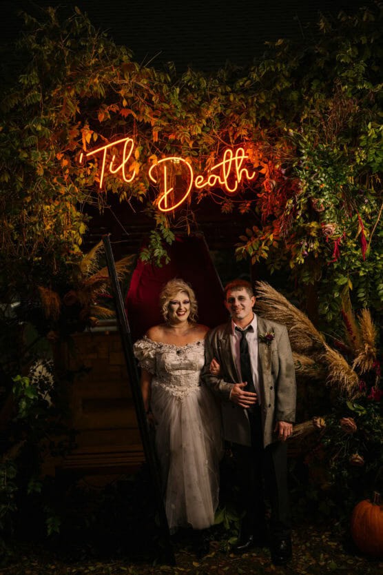A couple in spooky wedding attire poses under a neon sign reading Til Death, surrounded by lush, autumnal foliage and pampas grass, creating a moody, festive atmosphere. by Justin Salem Meyer
