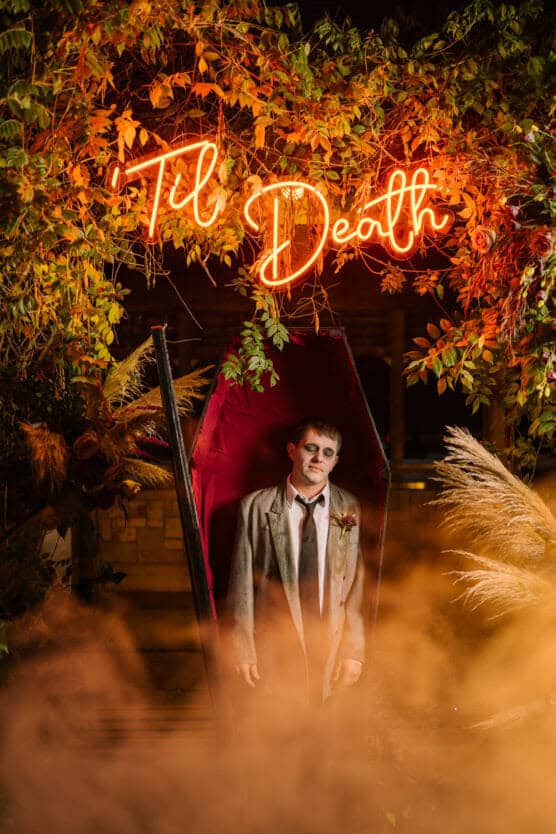 A person dressed as a zombie groom stands in an open coffin, surrounded by foliage and orange fog, with a neon sign above reading Til Death. by Justin Salem Meyer