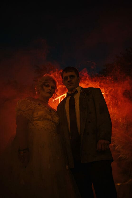 Two people in vintage formal wear stand close together, lit by eerie red and orange light and surrounded by swirling smoke, creating a dramatic, mysterious atmosphere at night. by Justin Salem Meyer