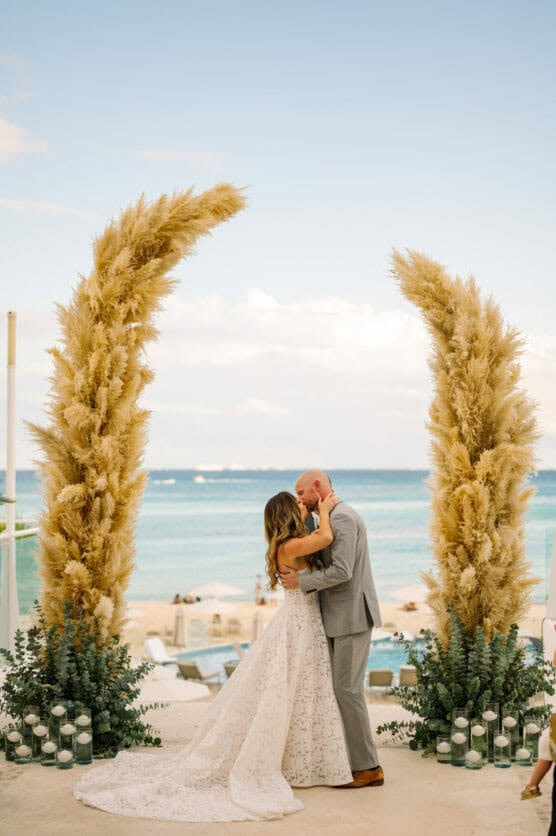 A bride and groom embrace and kiss at an outdoor beachside wedding ceremony, framed by two tall pampas grass arrangements, with the ocean and blue sky in the background. by Justin Salem Meyer