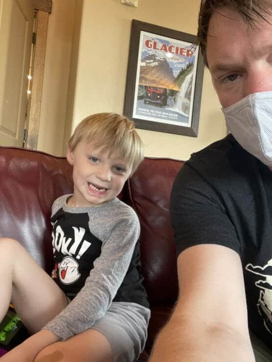 A young child with blonde hair sits on a red couch, smiling at the camera. An adult wearing a face mask is partially visible beside the child. In the background is a framed “Glacier” travel poster on the wall. by Justin Salem Meyer