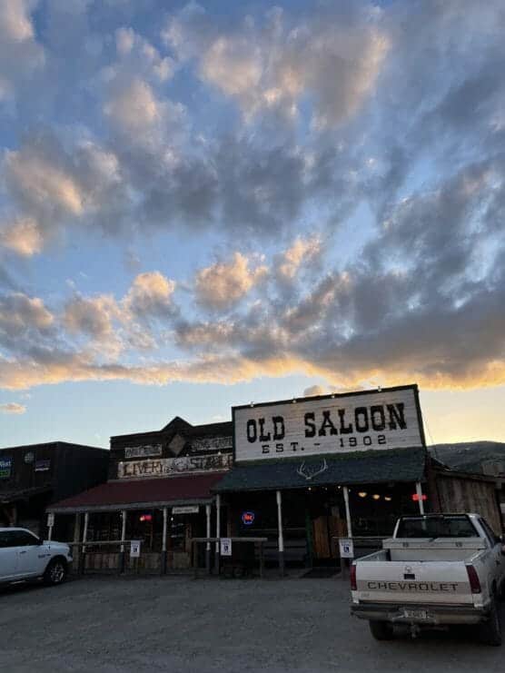 A rustic western-style building labeled Old Saloon Est. 1902 stands under a sunset sky with scattered clouds. Two pickup trucks are parked in front of the saloon and nearby wooden storefronts. by Justin Salem Meyer