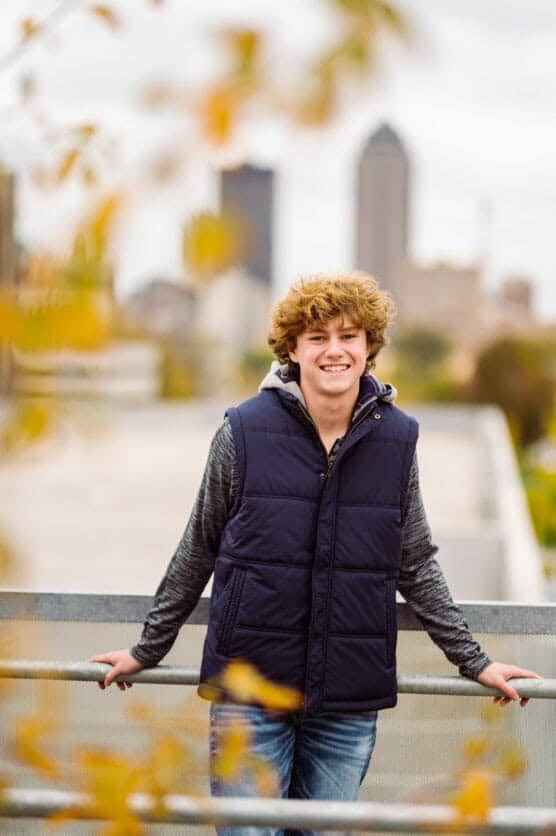 A smiling teenage boy with curly blond hair stands outdoors, leaning on a railing. He wears a navy blue puffer vest over a gray hoodie. Blurred autumn leaves and a city skyline appear in the background. by Justin Salem Meyer