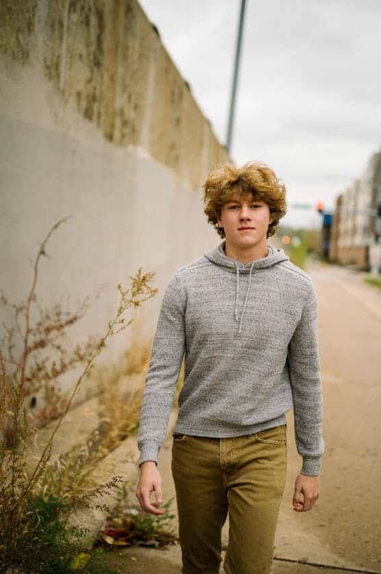 A young person with curly blond hair, wearing a gray hoodie and tan pants, walks along a city sidewalk beside a concrete wall on a cloudy day. by Justin Salem Meyer