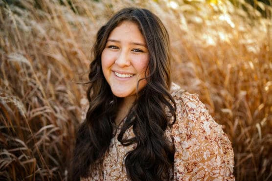 A young woman with long, dark wavy hair smiles warmly while standing in front of tall, golden-brown grasses. She wears a light brown floral-patterned top and looks directly at the camera. by Justin Salem Meyer