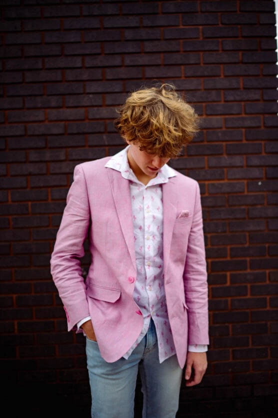 A young person with wavy blond hair wears a pink blazer over a white shirt with pink patterns and light blue jeans, standing against a dark brick wall while looking down with one hand in a pocket. by Justin Salem Meyer