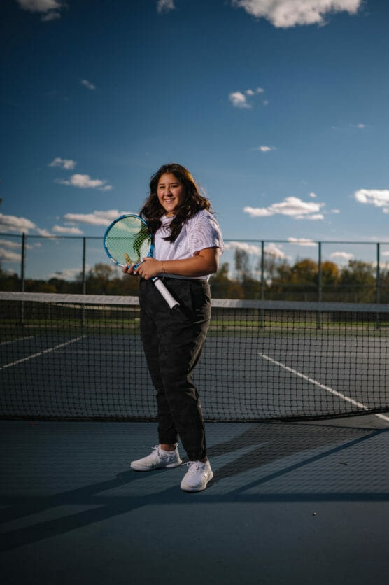 A woman stands on an outdoor tennis court in front of the net, holding a tennis racket and smiling. She is dressed casually in a white shirt, dark pants, and white sneakers. The sky is blue with scattered clouds. by Justin Salem Meyer