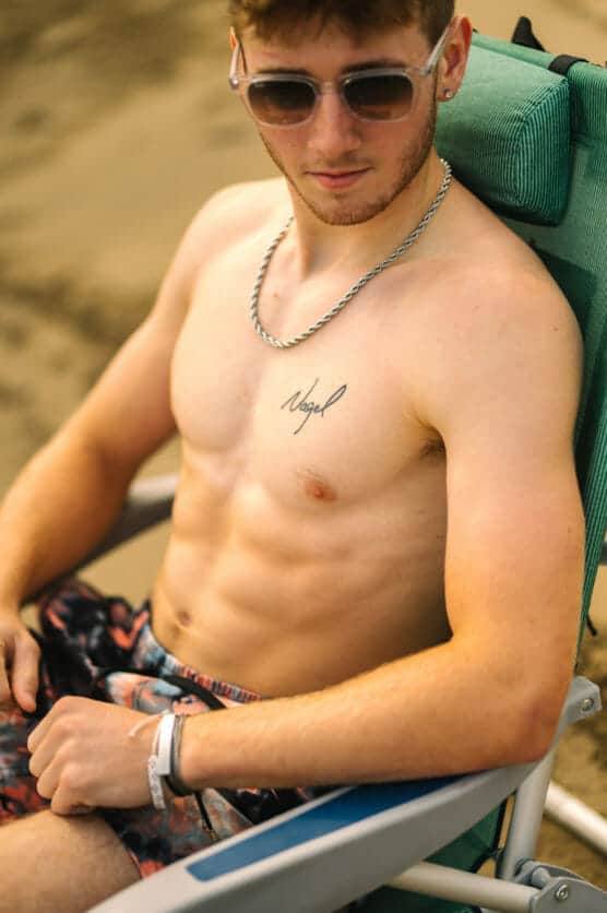 A young man wearing sunglasses and swim trunks relaxes shirtless on a green beach chair. He has a chain necklace, a wristband, and a tattoo that says Angel on his chest. The background is sandy. by Justin Salem Meyer