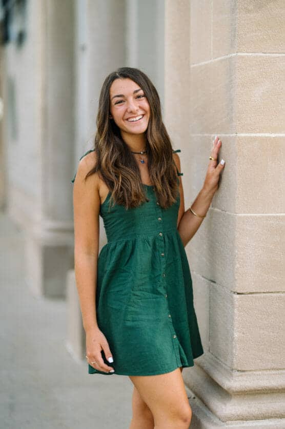 A young woman with long brown hair wearing a sleeveless green dress stands outdoors, smiling and leaning against a light-colored stone wall. by Justin Salem Meyer