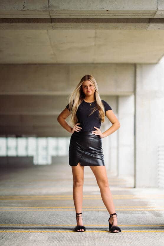 A young woman with long blonde hair stands confidently in a modern, concrete parking garage. She wears a black t-shirt, black leather mini skirt, and black heels, with her hands on her hips. by Justin Salem Meyer