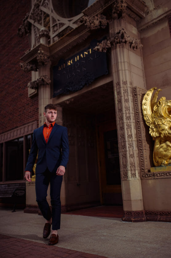 A man in a navy suit and orange shirt walks confidently on a sidewalk in front of an ornate building with a sign reading MERCHANTS BANK and a large gold decorative element. by Justin Salem Meyer