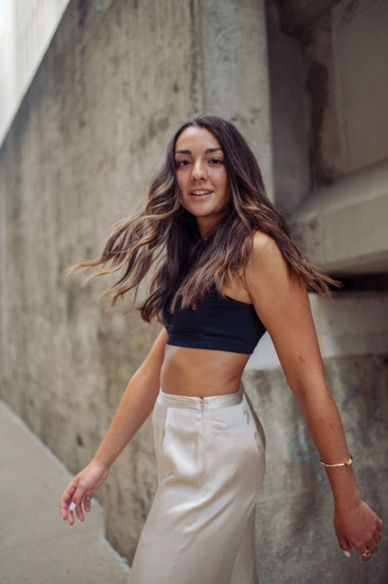A young woman with long brown hair wearing a black crop top and high-waisted cream pants walks along a concrete wall, looking at the camera with a slight smile. by Justin Salem Meyer