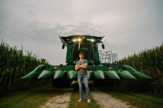 A young man stands with arms crossed in front of a large green combine harvester on a dirt path between tall cornfields under a cloudy sky. by Justin Salem Meyer