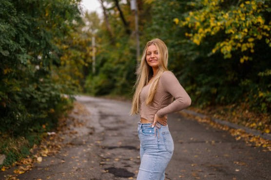 A young woman with long blonde hair, wearing a tan long-sleeve top and light blue jeans, stands on a tree-lined road and smiles at the camera. The background shows green and yellow foliage. by Justin Salem Meyer