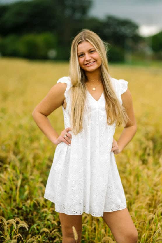 A young woman with long blonde hair stands in a field, wearing a sleeveless white dress and smiling with her hands on her hips. The background features tall grass and blurred green trees. by Justin Salem Meyer
