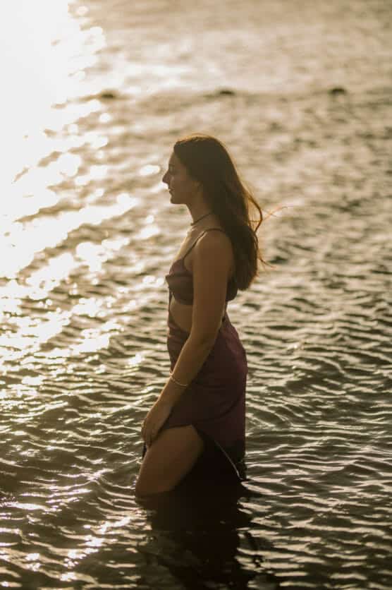 A woman in a sleeveless dress stands knee-deep in rippling water at sunset, with sunlight reflecting off the waters surface and her hair gently blowing in the breeze. by Justin Salem Meyer