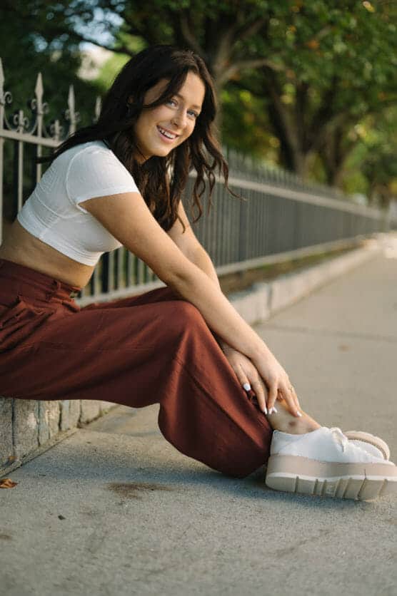 A young woman with long dark hair, wearing a white crop top, rust-colored pants, and white platform shoes, sits on a sidewalk near a black metal fence, smiling at the camera with trees in the background. by Justin Salem Meyer