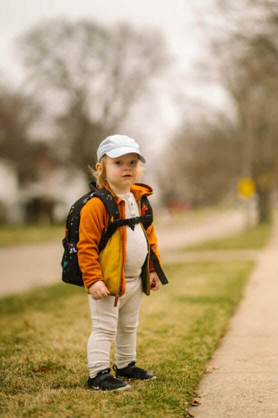 A young child wearing a light blue cap, orange jacket, white pants, and black shoes stands on a grassy area next to a sidewalk, carrying a black backpack. The background is blurred with trees and houses. by Justin Salem Meyer