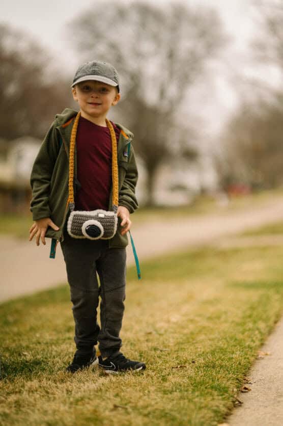A young child stands on a grassy area beside a sidewalk, wearing a green jacket, red shirt, gray pants, and a gray cap. The child has a toy camera around their neck and trees and houses are visible in the blurred background. by Justin Salem Meyer