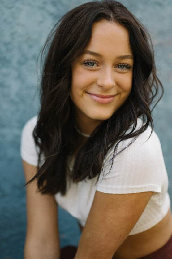A young woman with long, wavy dark hair and blue eyes smiles softly at the camera. She is wearing a white short-sleeve top and is posed against a textured blue background. by Justin Salem Meyer