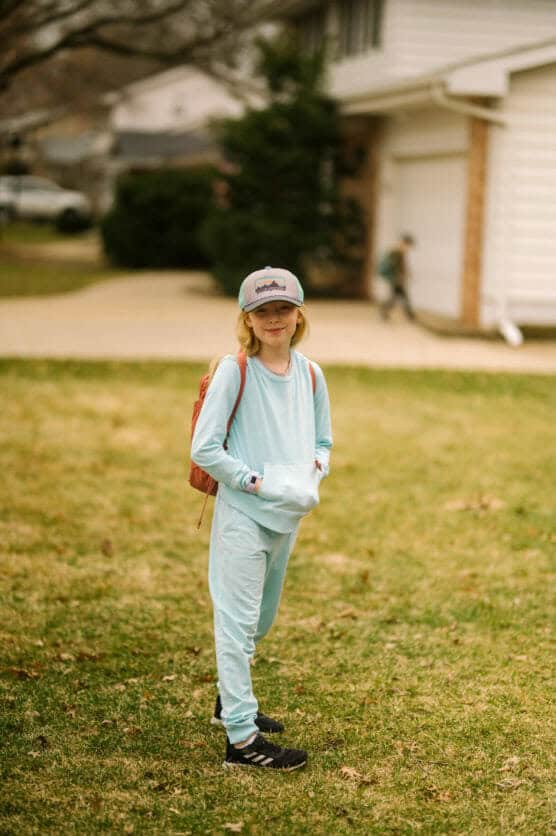 A child in a light blue outfit and cap stands on a grassy lawn with a backpack, smiling. A house and another child are visible in the blurred background. by Justin Salem Meyer