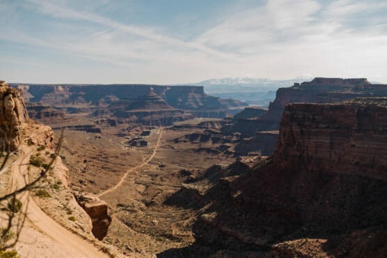 A wide canyon landscape with layered rock formations, a winding dirt road, and distant snow-capped mountains under a partly cloudy sky. by Justin Salem Meyer