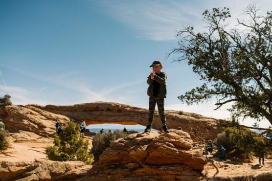 A young boy stands on a rocky formation in a desert landscape, wearing a hat and sunglasses, with a natural stone arch and several people in the background under a clear blue sky. by Justin Salem Meyer